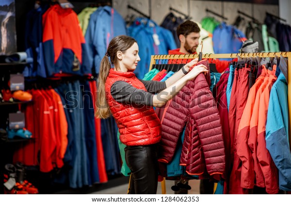 Young woman choosing winter clothes\
picking up down jackets on the hanger in the sports\
shop