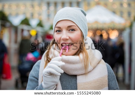 Young woman in Christmas market eating candy cane wearing knitted warm hat and scarf. Illuminated and decorated fair kiosks, shops and carousel on background. Helsinki, Finland