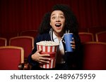 A young woman in a cinema watches a movie, holding a bucket of popcorn and a drink, reacting with excitement and surprise.