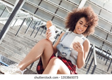 Young Woman In The City Street Sitting On Concrete Stairs Holding Sandwich Checking Social Media On Smartphone Smiling Cheerful