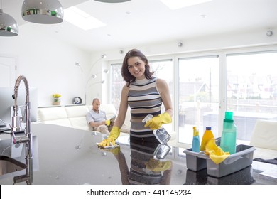 A Young Woman Is Cleaning A Counter Top. She Is Looking At The Camera Smiling While Her Partner Is Sat On The Couch Reading.