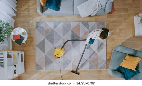 Young Woman Cleaning A Cozy Living Room With A Vacuum Cleaner. Modern Interior With Carpet, Sofa, Chair, Coffee Table, Shelf, Plant And Wooden Floor. Top Down.