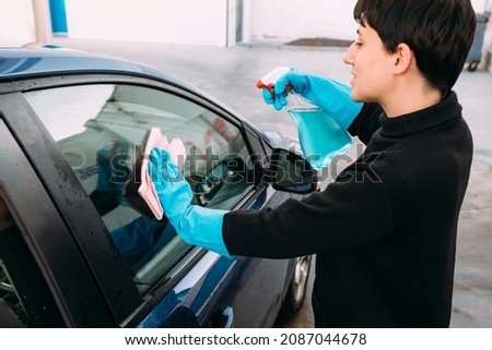 Young woman cleaning the window of a car using a spray and a pink cloth while wearing blue rubber gloves. Concept of self-service car washing. 