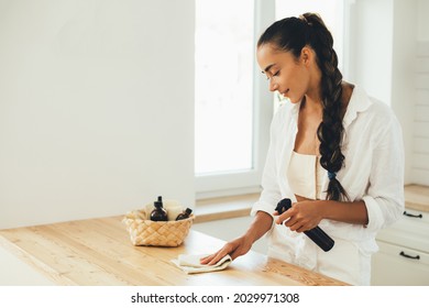 Young woman cleaning wooden table using spray in glass bottle and natural rag in a kitchen.