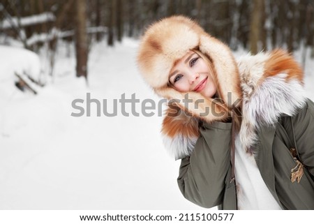 Young woman close up portrait at winter nature background, wearing parka jacket, fur hat and bright leather gloves, healthy activity. Blonde girl with blue eyes and beautiful smile