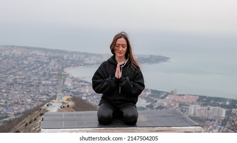 A young woman with closed eyes, long hair, sits on top of a mountain with her hands clasped in prayer.