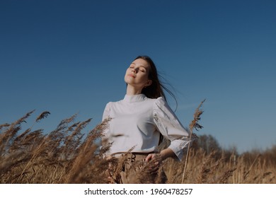 Young woman with closed eyes in white blouse standing in field of dry pampas grass in front of blue sky and sun. Urban style and street fashion. Girl in casual outfit looking up in the sky - Powered by Shutterstock