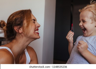 Young Woman Closing Eyes With Laughing Little Boy Putting On Makeup On Her Lips. Small Kid Holding A Lipstick And Laughing While Doing Makeup To His Mother.