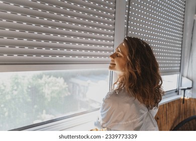 Young woman closing roller shutters on the balcony on a summer day - Shutterstock ID 2339773339