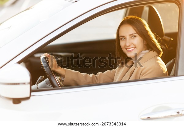 A young woman in coat is driving a car. Smiling\
female looks at camera and smiles in warm natural light. Taking\
driving courses