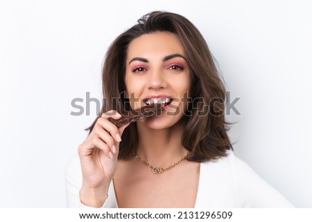 Young woman in a cocktail dress, gold chain, bright spring pink makeup on a white background. Holds a healthy cereal chocolate delicious bar.
