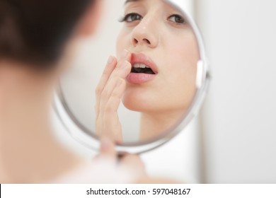 Young Woman With Cold Sore Looking In Mirror At Home