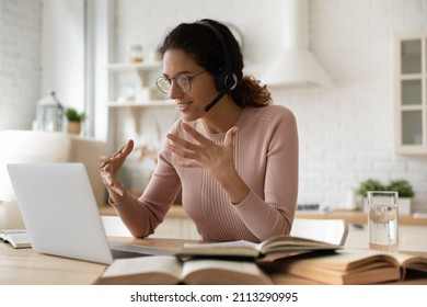 Young woman college student e-learn sit in kitchen wear headset look at laptop talk to tutor use videoconference, heap of textbooks on table. Digital online educational course, videocall event concept