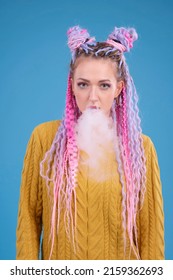 Young Woman With Colored Colorful Hair Smoking Electronic Cigarette, Blowing Smoke From Mouth, Studio Portrait. Bright Woman With Afrobraids. Blue Solid Background. Arrogant Person