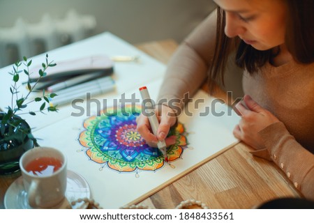Young woman colouring mandala with markers and white rosary on table with cup of coffee near flower vase at home. Art therapy and meditation concept. Hobby and leisure during quarantine. Wellbeing.
