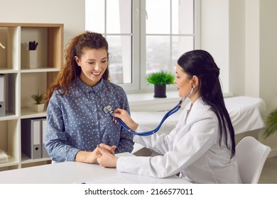 Young woman comes to clinic for heart and lungs checkup. Friendly female doctor sitting at desk in modern medical office, holding stethoscope, listening to patient's breath or heartbeat and smiling - Powered by Shutterstock