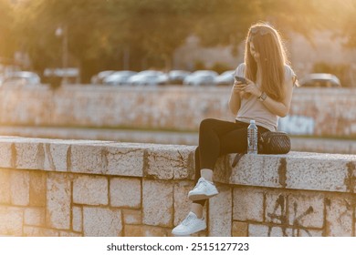 A young woman is comfortably seated on a stone wall near a river, focused on her smartphone. The golden hour sunlight casts a warm glow, enhancing the tranquil atmosphere. - Powered by Shutterstock