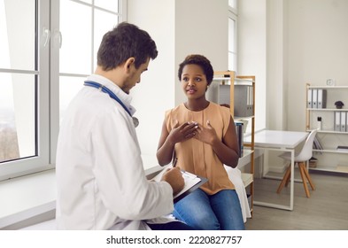 Young Woman Complains During Medical Examination Of Chest Pain And Difficulty Breathing. Young Doctor Records Complaints Of Dark-skinned Patient. Heart Attack And Cardiovascular Disease Care Concept.