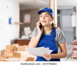 Young Woman Construction Worker Standing In Construction Site, Holding Documents And Talking On Phone.