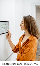 Young Woman Controlling Home With A Digital Touch Screen Panel Installed On The Wall In The Living Room. Concept Of A Smart Home And Mobile Application For Managing Smart Devices At Home