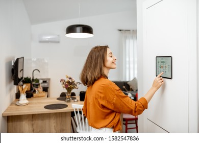 Young Woman Controlling Home With A Digital Touch Screen Panel Installed On The Wall In The Living Room. Concept Of A Smart Home And Mobile Application For Managing Smart Devices At Home