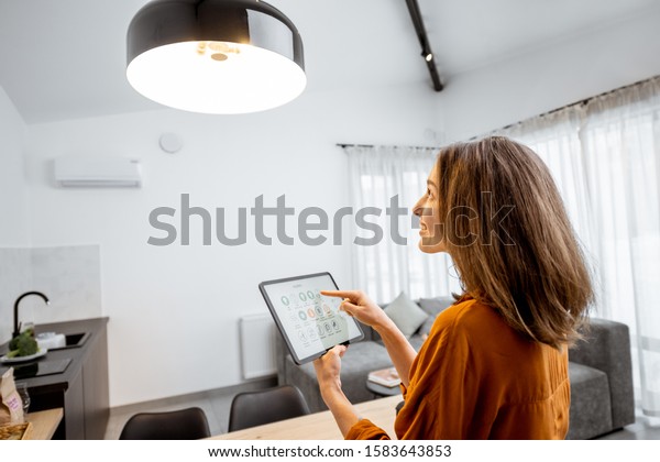 Young woman controlling home light with a digital\
tablet in the living room. Concept of a smart home and light\
control with mobile\
devices