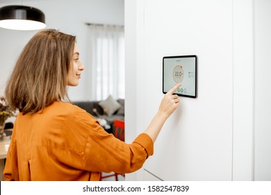 Young Woman Controlling Temperature In The Living Room With A Digital Touch Screen Panel Installed On The Wall. Concept Of Heating Control In A Smart Home