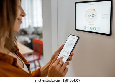 Young woman controlling temperature in the living room with smart phone and digital touch screen panel. Concept of heating control in a smart home