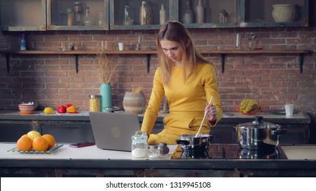 Young woman cooking dinner and reading recipe. Adult lady using internet on pc in the kitchen. Caucasian model sitting near cooker looking on the laptop screen. Happy girl typing on keyboard in loft