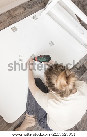 Young woman with cordless screwdriver assembling white folding table on the floor