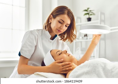 Young woman cosmetologist or dermatologist directing lamp for facial treatment to lying young woman in beauty spa salon. Facial treatment, massage, skincare, cosmetology concept