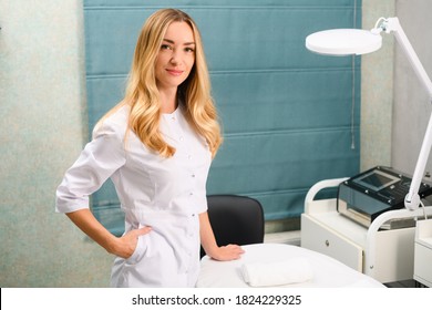 Young woman, cosmetology doctor in white uniform posing at beauty clinic cabinet with couch, table lamp and apparatus for skin treatment procedures
