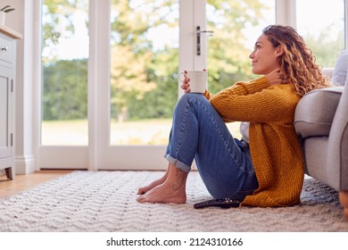 Young Woman In Cosy Warm Jumper Sitting On Floor At Home Looking Out Of Window With Hot Drink