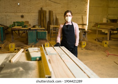 Young Woman Covered In Sawdust Inside A Woodshop. Female Carpenter Standing Next To A Table Saw Machine And Wearing Security Glasses, Gloves And A Face Mask