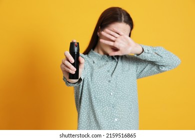 Young woman covering eyes with hand and using pepper spray on yellow background