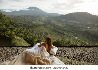 Young woman covering a white blanket sit in hammock  at the view of nature of mountains with green jungle. travel in the morning time with mountain view and natural green view feeling chill and relax.