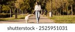 A young woman in cozy autumn attire walks her dog through a vibrant park surrounded by trees.