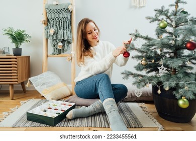 Young woman in cozy sweater decorating potted Christmas tree with small glass baubles in light modern Scandinavian interior. Eco-friendly winter holidays. Christmas tree in a pot. Selective focus