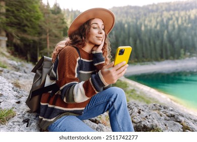 A young woman in a cozy sweater and hat sits on a rock by a beautiful lake, smiling and using her phone. Adventure, nature, blogging. - Powered by Shutterstock