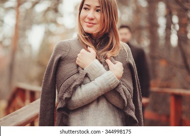 Young Woman In Cozy Warm Cardigan Walking Outdoor In Autumn Forest, With Boyfriend On Background