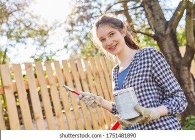 Young Woman As A Craftsman Trainee With Paint Can For Painting Wooden Fence In The Garden