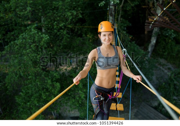 Young woman crossing the chasm on the rope\
bridge. Sports entertainment in the\
park.