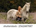 A young woman cuddle and interact with her white spanish horese on a wildflower field in autumn outdoors