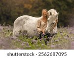 A young woman cuddle and interact with her white spanish horese on a wildflower field in autumn outdoors