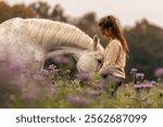 A young woman cuddle and interact with her white spanish horese on a wildflower field in autumn outdoors