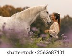 A young woman cuddle and interact with her white spanish horese on a wildflower field in autumn outdoors