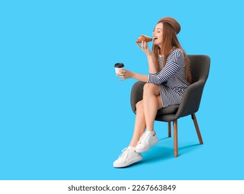 Young woman with cup of coffee eating croissant in armchair on blue background