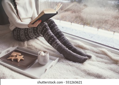 Young Woman With Cup Of Coffee And Gingerbread Is Reading A Book Sitting Home Relaxing By The Window. Snow Winter Home Chill Out Concept