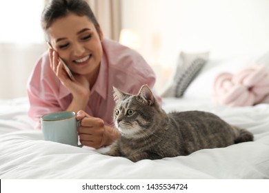 Young Woman With Cup Of Coffee Talking On Phone While Lying Near Cute Cat In Bedroom. Pet And Owner