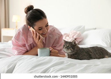 Young Woman With Cup Of Coffee Talking On Phone While Lying Near Cute Cat In Bedroom. Pet And Owner